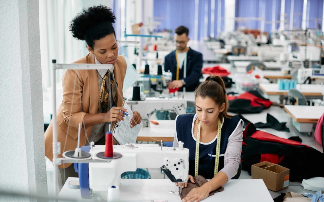 Young seamstress sewing clothes while production line manager is overseeing her.